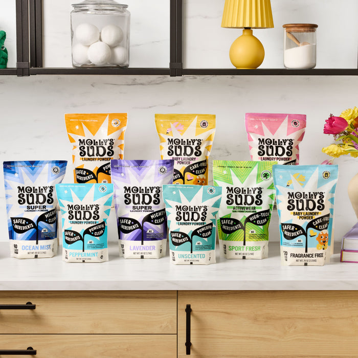 Ten vibrant bags of Molly’s Suds, including Activewear Laundry Powder, sit on a kitchen counter with flowers, a yellow lamp, and stacked jars in the background.