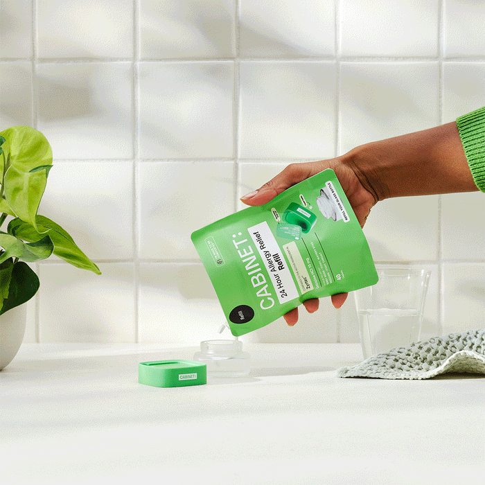 A hand pours green dishwasher detergent from a Cabinet Health pouch into a dishwasher compartment, set against eco-friendly white tiles, a leafy plant, and a countertop with a towel and glass.