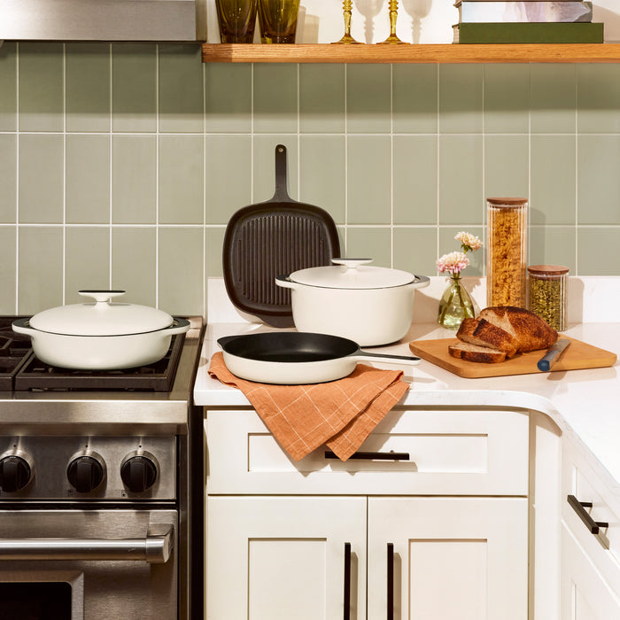 In the kitchen scene, a stovetop features a white pot and grill pan. On the countertop, a Caraway Cast Iron Braiser with Lid, 3.5 qt offers superior heat retention beside a white Dutch oven and skillet on an orange towel. Sliced bread sits on a board with a knife near pasta and grain containers.