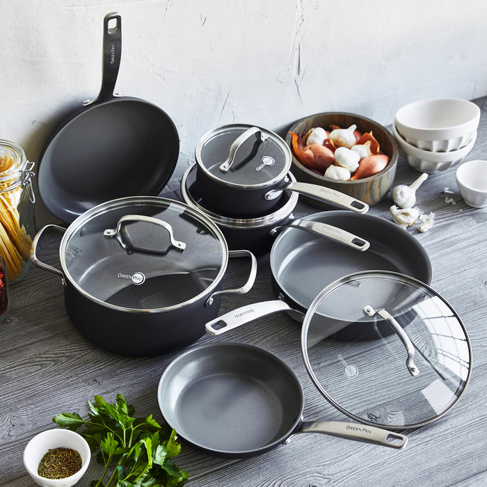The GreenPan Chatham Ceramic 10-Piece PFAS-Free Non-Stick Cookware Set, featuring black pots, pans, and lids with silver handles and Thermolon surfaces, is displayed on a gray wooden surface. Nearby are white bowls, pasta jar, garlic, shallots, fresh herbs, against a light-colored wall backdrop.