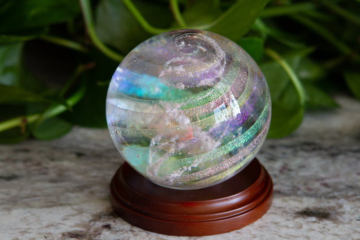 Dichroic rainbow orb on a circular wooden stand with a leafy green backdrop