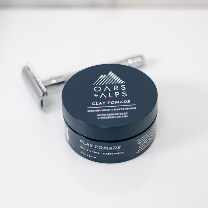 A round Oars + Alps Clay Pomade container sits on a white surface beside a silver razor. The label features its clay pomade formula for medium matte hold, frizz-free style, and kaolin clay.