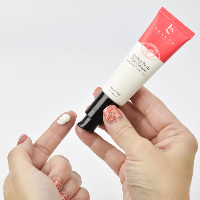 A person applies Coffee Bean Awakening Eye Cream from Beauty by Earth onto their finger. The tube’s cap is off, showcasing its coffee seed extract. Their nails gleam with glittery red polish as they aim to diminish dark circles; the tube displays a white and red design.