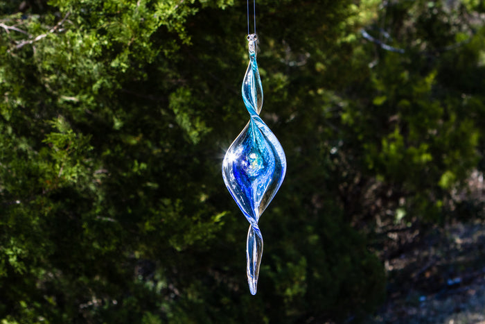 Dichroic spiral suncatcher infused with cremains elongated blue and clear glass pendant hanging outdoors.