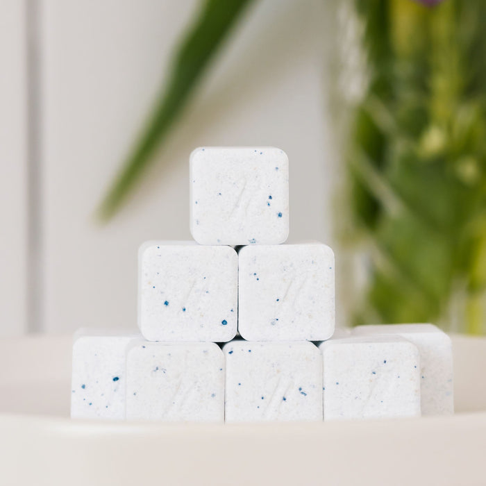 Seven white Dishwasher Detergent Cubes with blue specks from Nellie's are stacked in a bowl, set against a softly blurred green and purple background that evokes planet-friendly, natural clean living.