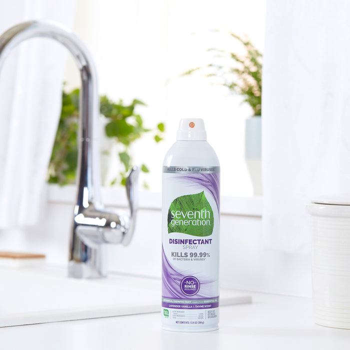A Seventh Generation Disinfectant Spray can with a green leaf design label sits on a white kitchen countertop near the sink, highlighting its germ-killing power on nonporous surfaces. Sunlight illuminates potted plants in the background from a nearby window.