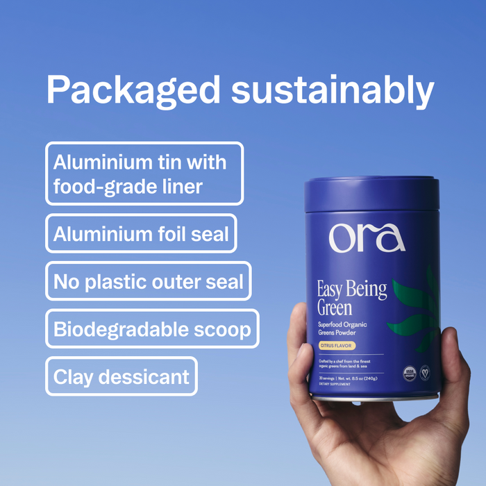 A hand holds a blue Ora Organic Easy Being Green: Daily Active Superfood Greens Powder tin with organic prebiotics and adaptogens, set against a clear sky. Text highlights its sustainable packaging—aluminum tin, foil seal, biodegradable scoop, no plastic.