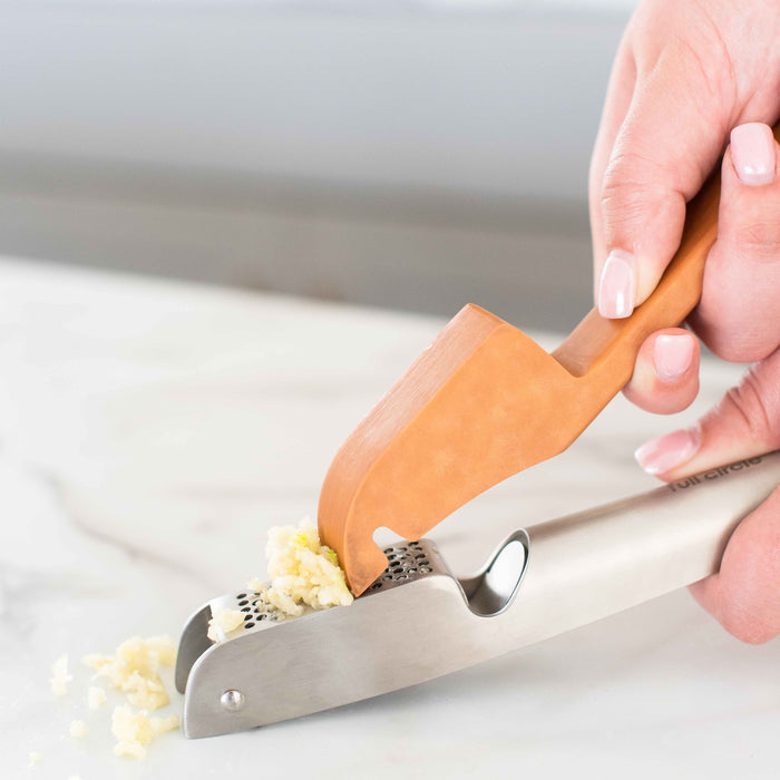 A hand grips the wooden handle of Full Circles Easy-Clean Garlic Press, scraping crushed garlic from the stainless steel, dishwasher-safe press and spreading it over a white marble surface.