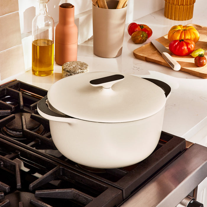 A Caraway Cast Iron Dutch Oven with Flat Lid, 6.5 qt, sits on a modern stovetop surrounded by oil bottles, a jar of utensils, and a cutting board with tomatoes—perfect companions for this pot renowned for excellent heat retention.