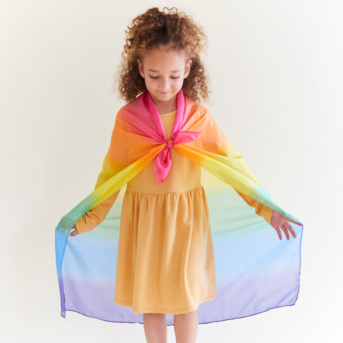 A young girl with curly hair twirls in a yellow dress and a colorful Enchanted Playsilk cape from Sarahs Silks, made of mulberry silk and tied around her neck. She smiles widely, embracing open-ended play as she holds the vibrant cape with outstretched arms against a plain background.