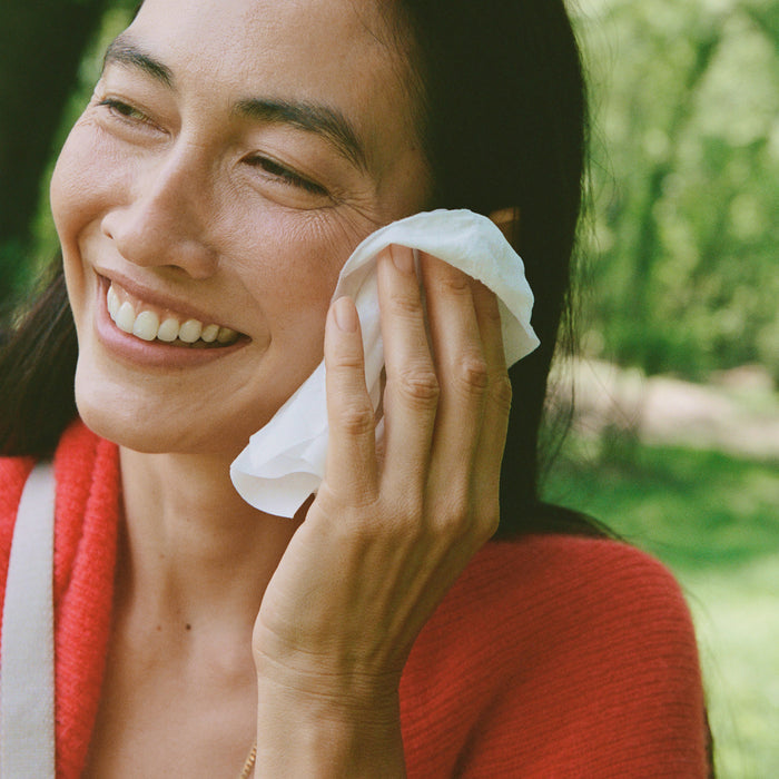 A woman in a red sweater with long dark hair smiles as she uses an individually wrapped Ursa Major Essential Face Wipe outdoors, surrounded by greenery.