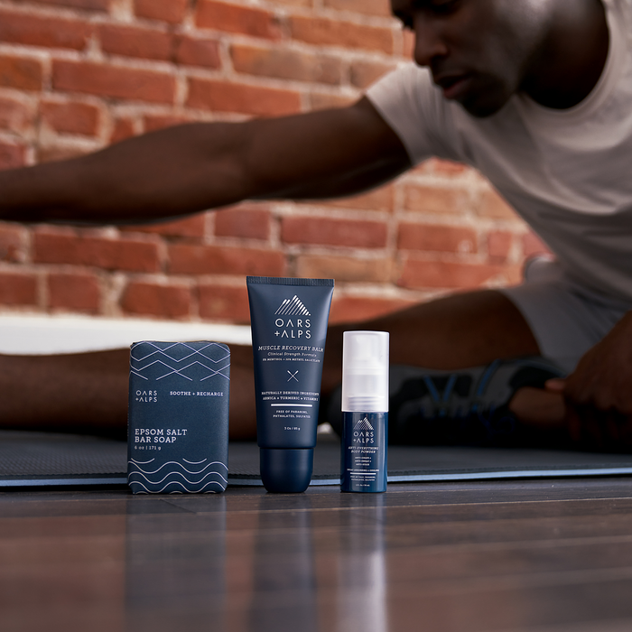 A man is stretching on a yoga mat by a brick wall, concentrating on three Oars + Alps items: Epsom Salt Bar Soap, Muscle Recovery Balm, and an Anti-Everything Body Powder, all neatly arranged in the foreground.