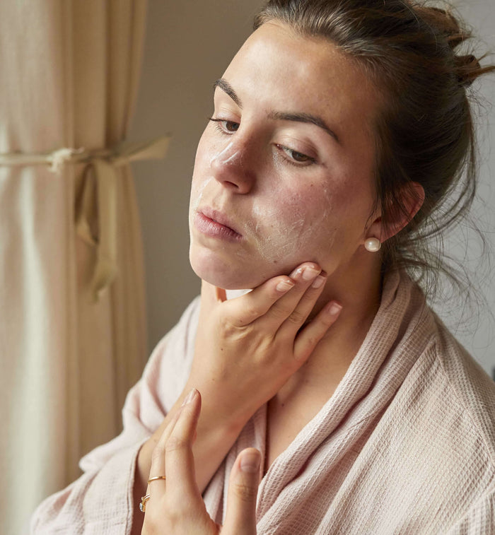 A person in a light pink robe applies UpCircles Face Moisturizer with Vitamin E + Aloe Vera while looking into a mirror, gently touching their cheek and neck. Beige curtains with fabric ties are seen in the background.