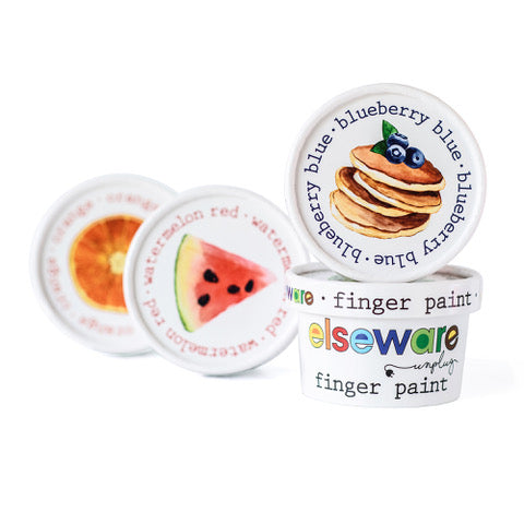 Four small tubs of eco-kids Finger Paint Set are shown. The front tub has a white label with colorful text, and the blurred background displays labels illustrated with a blueberry stack, watermelon slice, and orange slice. These finger paints are all-natural and food safe.