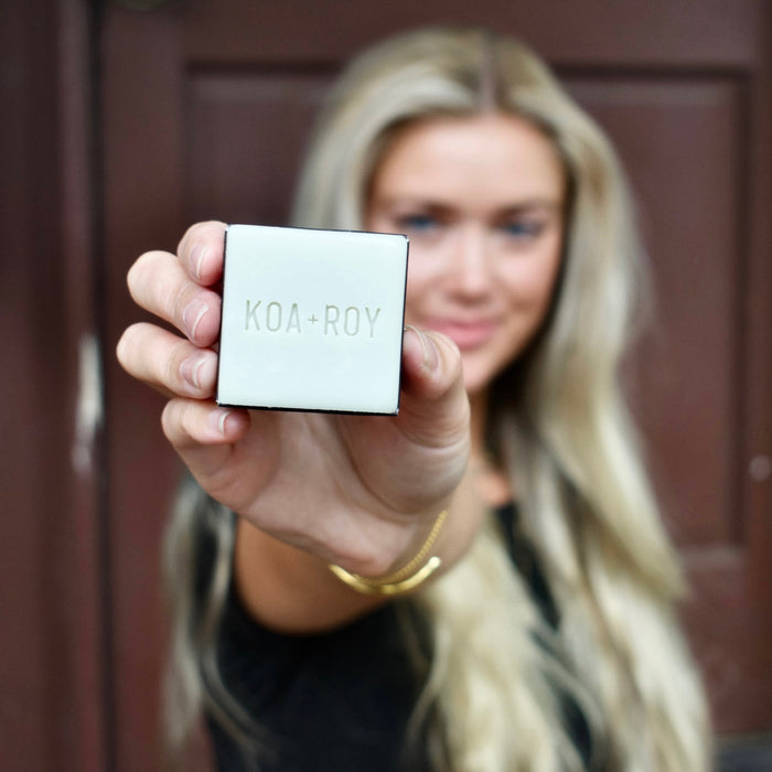 A woman with long blonde hair holds a small white KOA + ROY Fitness Cube box toward the camera, showcasing its plant-based muscle recovery benefits, with her face and the background slightly blurred.