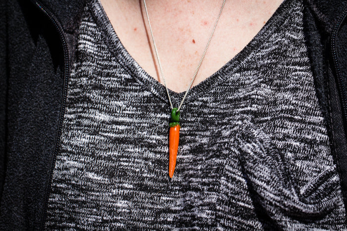 Orange glass carrot pendant with cremation ash and green top on a silver chain worn around the neck against a gray patterned sweater