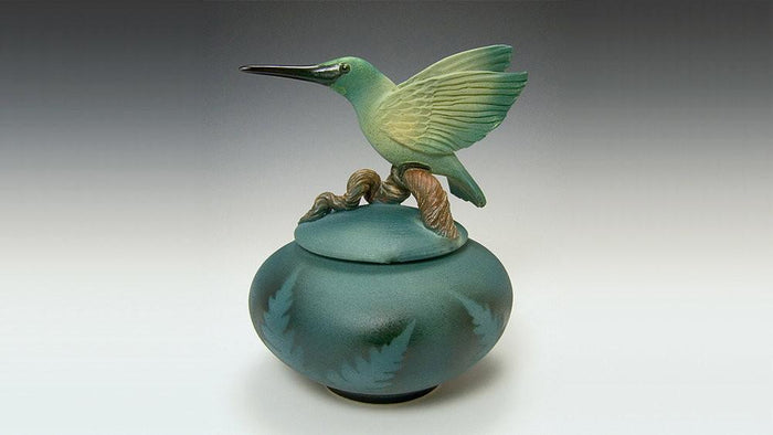 Teal round ceramic urn topped by a hummingbird figurine perched on a branch.
