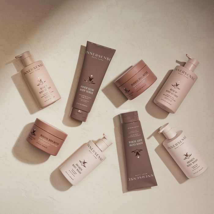 A flat lay of Innersense Organic Beauty, featuring the Inner Glow Body Scrub and other hair and body care products on a light surface. Neutral-tone bottles and tubes are arranged randomly, casting soft shadows. Perfect for achieving healthy skin.