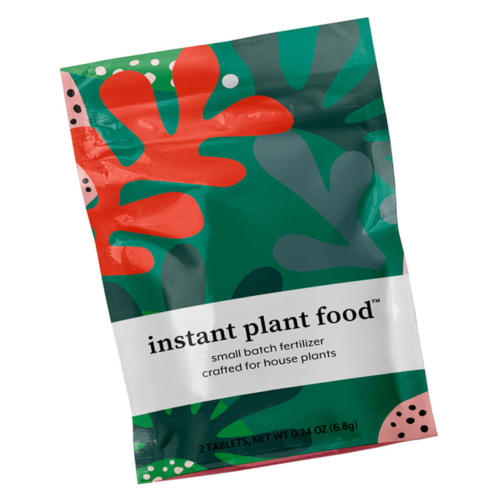 A colorful Instant Plant Food packet displays abstract red and green leaf designs. The label states its a small batch fertilizer for indoor plants, offering essential nutrients. It contains two tablets with a net weight of 0.24 oz (6.8g), perfect for your plants needs.