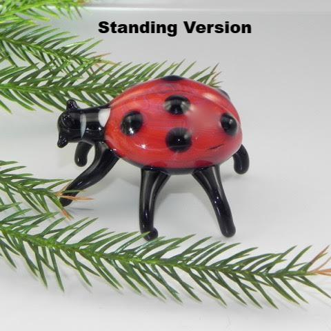 Standing version red glass ladybug cremains figurine with black legs on a white surface among pine needles