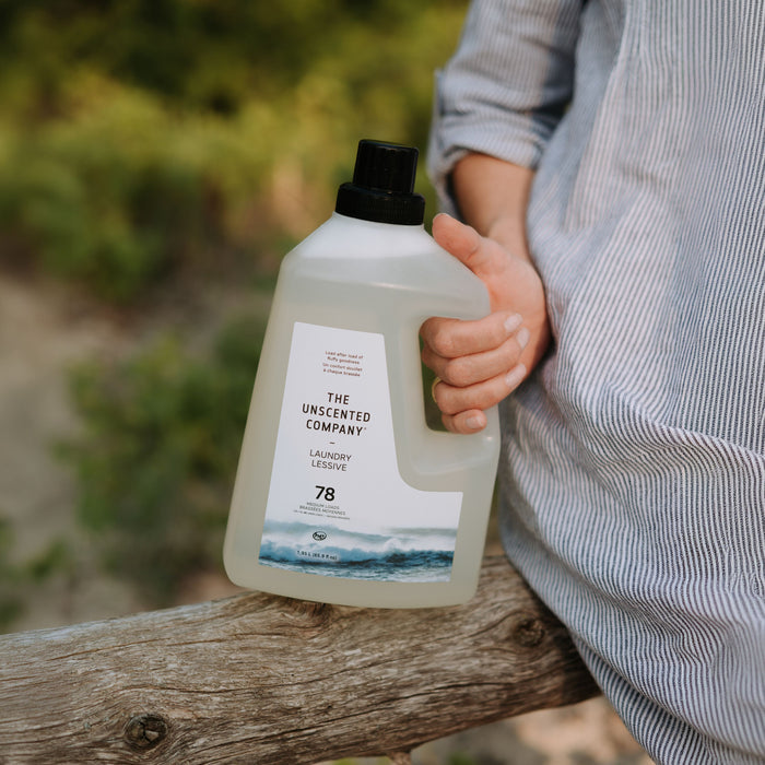 A person in a striped shirt holds a bottle of The Unscented Company Laundry Detergent outside amid green plants.