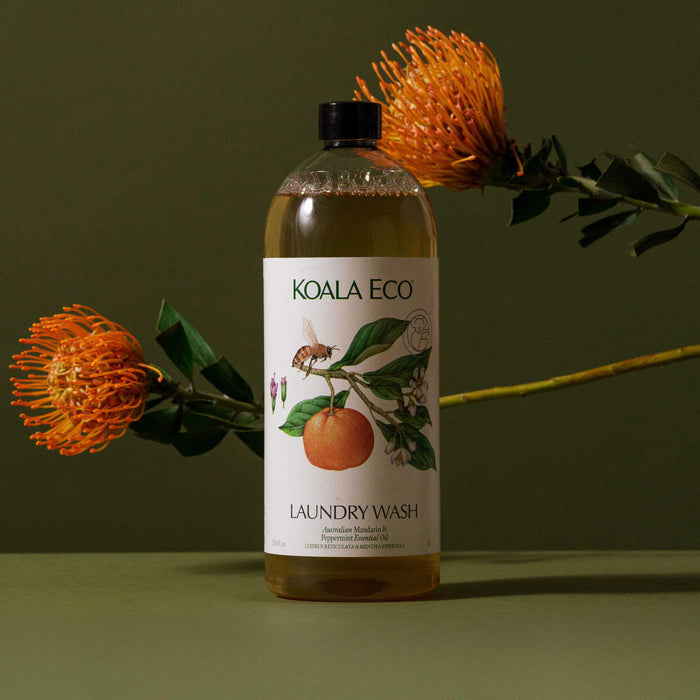 A bottle of Koala Eco Laundry Wash - Mandarin & Peppermint, featuring an orange and botanical illustrations on the label, is set against a green background with orange flowers.