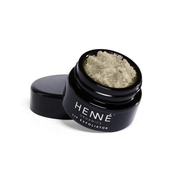 A black jar of Henné Organics Lip Exfoliator, with its lid removed and placed elegantly beside it, reveals a coarse, pale exfoliating formula crafted from organic ingredients and sugar crystals.