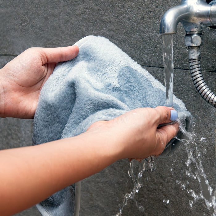 Hands wring out Tree To Tubs light blue Makeup Remover Towel under a metal faucet, set against a textured gray background.