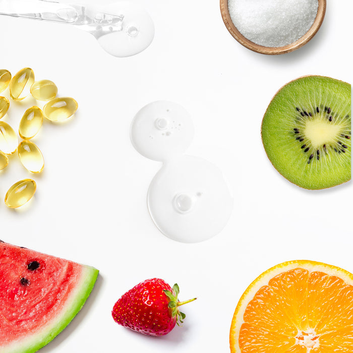 A white background displays a scattered array featuring vitamin capsules, fruits like strawberry, kiwi, orange, and watermelon slices, alongside gel-like drops. A bowl of sugar symbolizes the gentle exfoliating effect of Tree To Tubs Mandelic Acid 10% Gentle Exfoliating AHA Serum.