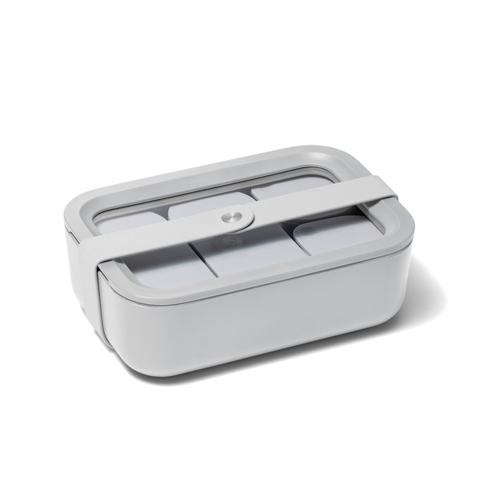 The Caraway Medium Glass Food Container has a grey rectangular design with a ceramic-coated transparent glass lid, secured by a white silicone strap. Featuring rounded edges and visible compartments, it offers a non-toxic, modern flair for stylish meal storage.