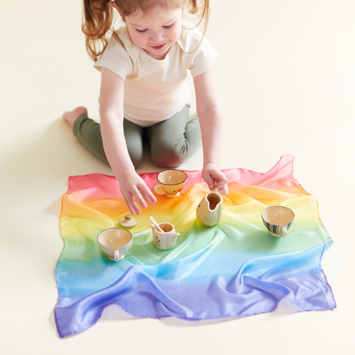 A young girl in a white shirt and green pants sits on the floor, engaged in open-ended play with small cups and a jug on Mini Playsilk for Open-Ended, Imaginative Play by Sarahs Silks. She is focused and smiling.