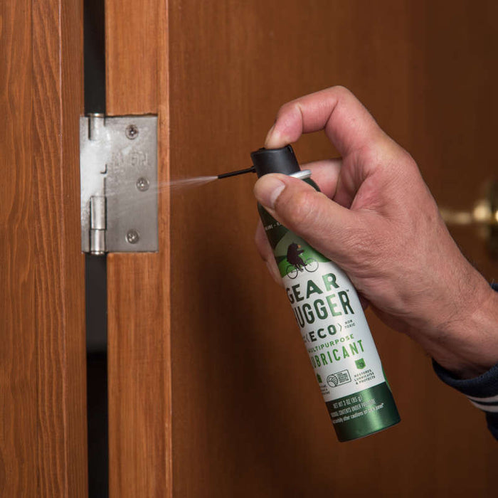 A hand sprays Gear Hugger Multi-Purpose Lubricant from a green and white can onto a door hinge, providing non-toxic rust protection as it is applied to the metallic hinge pin for maintenance.