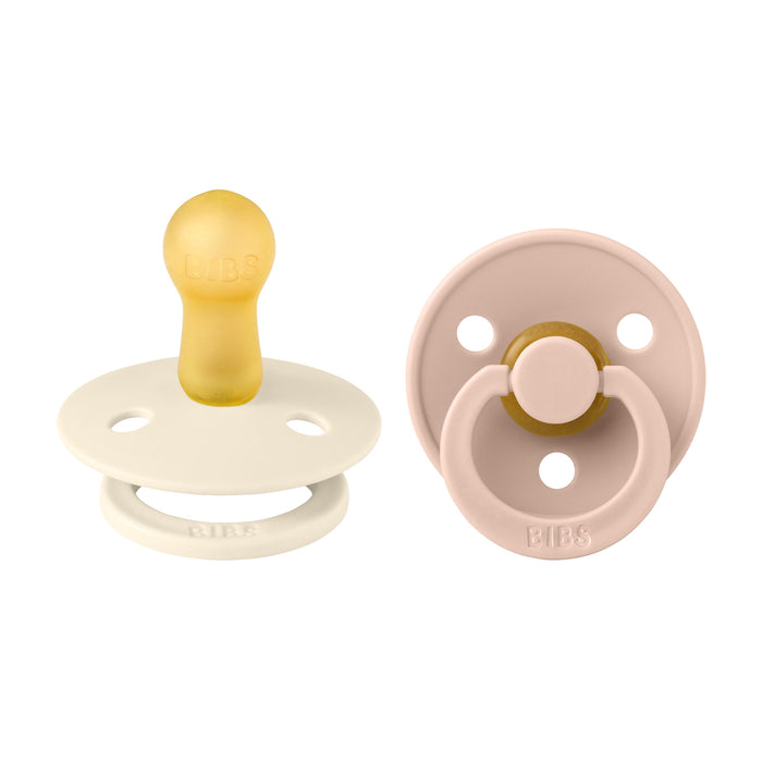 Two BIBS Natural Latex Rubber Pacifiers, Size 2 (6-18 Months), are shown. The left has a cream matte finish, while the BPA-free right one is light pink with an attached clip. Both have BIBS embossed on their bases for trusted breastfeeding support.