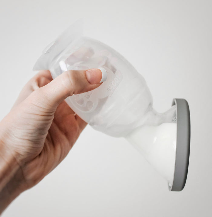 A hand holds a Haakaa New Mom Starter Pack translucent container with white liquid, likely milk. Its flexible, curved design and soft gray base suggest its perfect for breastfeeding needs like a silicone breast pump or milk storage bag.