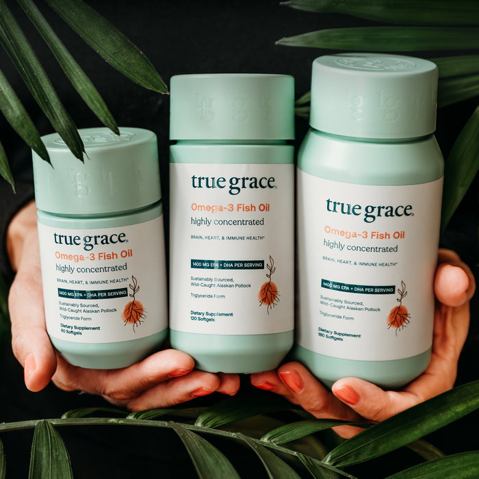 Three hands hold three green bottles of True Grace Omega-3 Fish Oil, surrounded by tropical leaves. The label highlights benefits for brain, heart, and immune health.