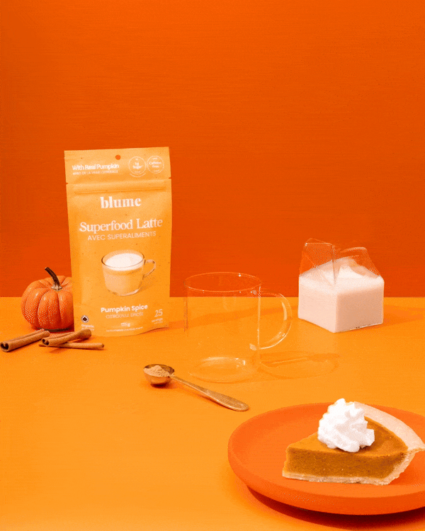 An orange-themed setup features a blume Organic Pumpkin Spice Latte Powder packet, highlighting its organic pumpkin flavor. The scene is completed with a clear mug, sugar container, cinnamon sticks, a mini pumpkin, a spoon of latte powder, and whipped cream-topped pumpkin pie on an orange plate.