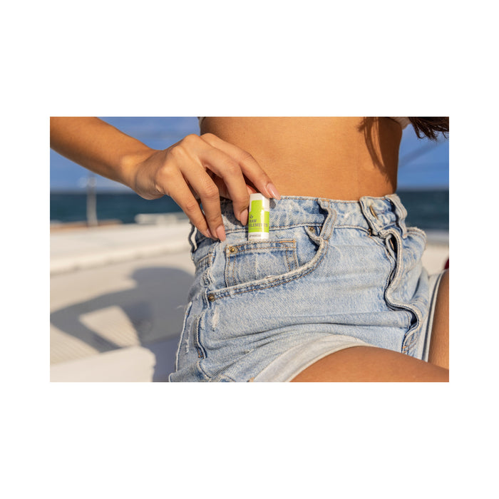 On a boat, against a serene ocean backdrop, someone in denim shorts slips the small green and white Outdoor Lip Rescue - SPF 30 by Raw Elements, offering broad spectrum protection, into their pocket.