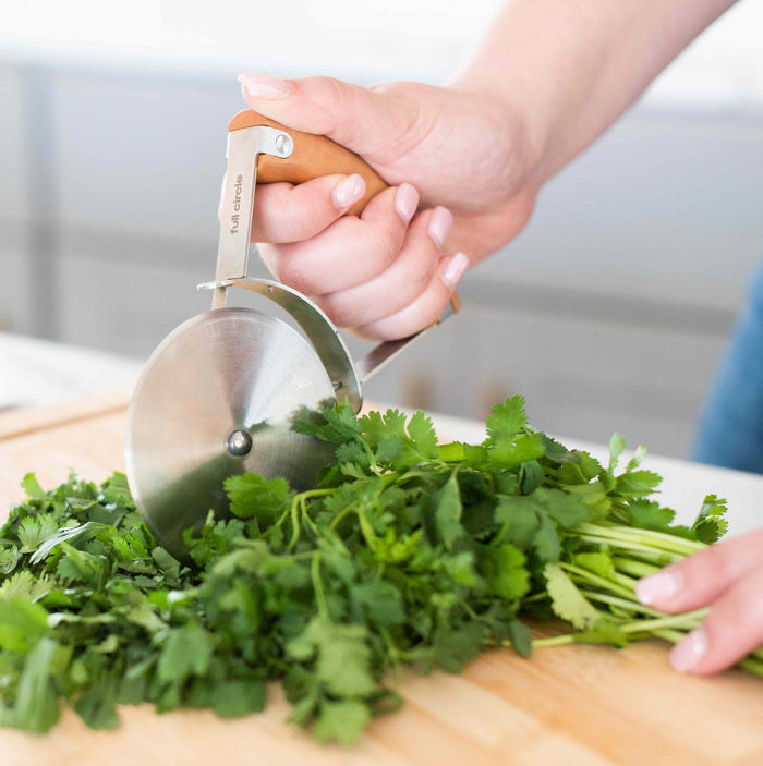 With Full Circles Pizza Wheel, featuring a hover-style handle, a person effortlessly achieves precision cutting as they chop fresh cilantro on a wooden cutting board.