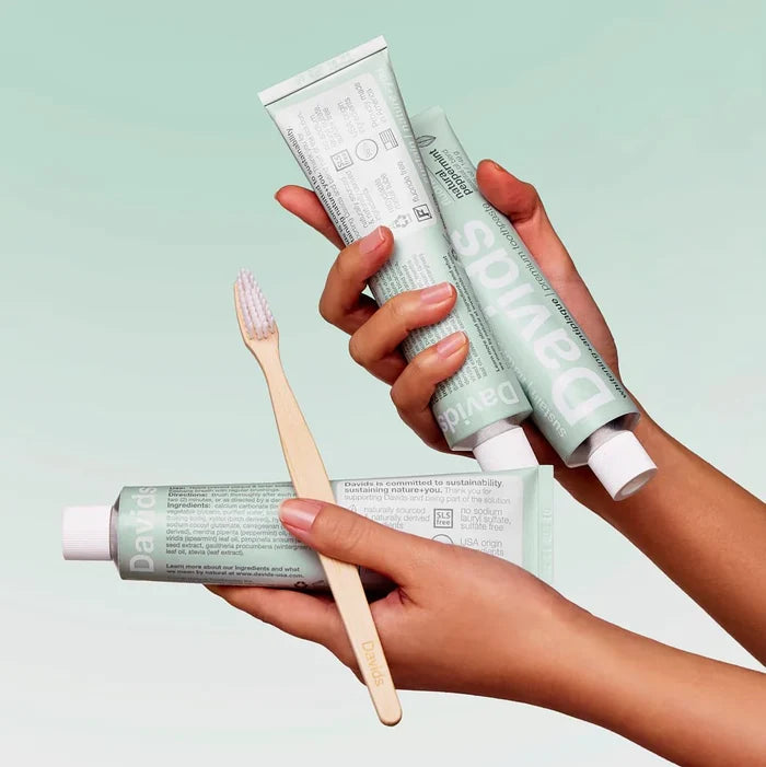 Three hands hold green tubes of Davids Premium Toothpaste – Whitening + Antiplaque and a wooden toothbrush with white bristles against a light green gradient background. Proudly EWG Verified, this fluoride-free toothpaste by Davids uses clean ingredients to deliver effective whitening and antiplaque benefits for a healthier smile.