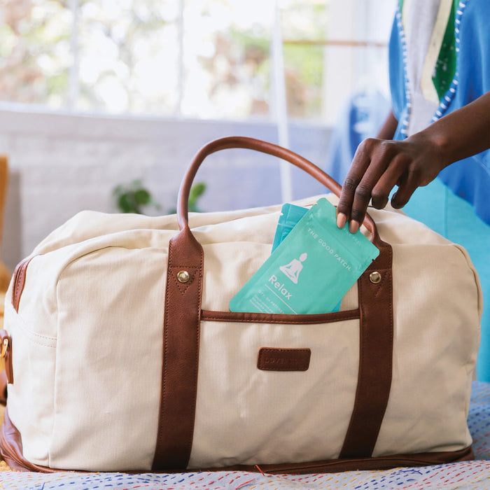 A person in a blue outfit gently places a teal package labeled Relax Plant Patch by The Good Patch into a beige canvas and brown leather duffel bag. The indoor scene is softly lit, evoking tranquility and stress relief.