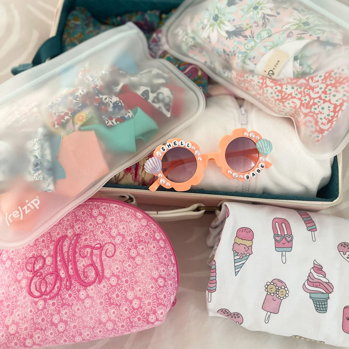An open suitcase packed with colorful clothes, a floral monogrammed pouch, floral hair bows, candy-adorned pink sunglasses, an ice cream and popsicle print shirt, and (re)zip Reusable Slim Deli & Leftover Bags for travel essentials.