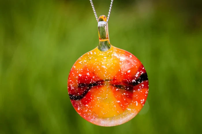 Round glass pendant resembling a setting sun in warm orange yellow and red tones with white specks on a silver chain