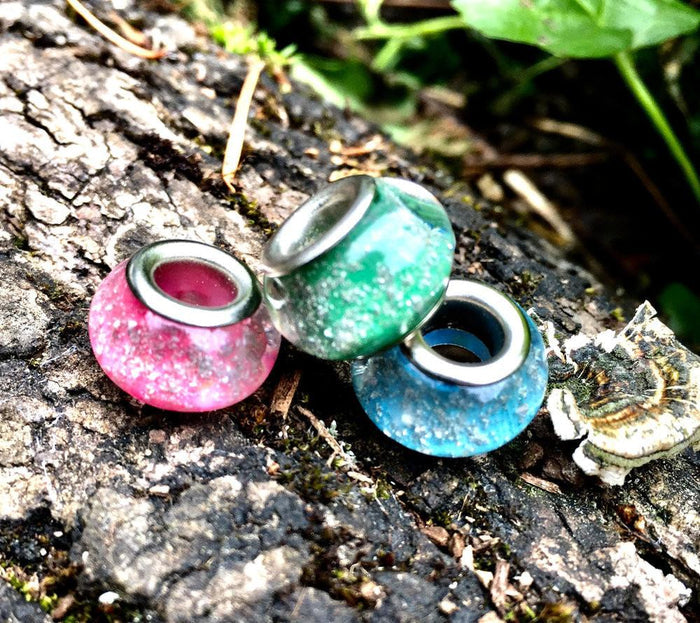 Pink, green, and blue speckled cremains beads arranged together on a mossy log.