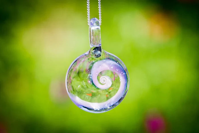 Purple toned glass spiral pendant infused with ashes, suspended from a silver chain against a green garden background