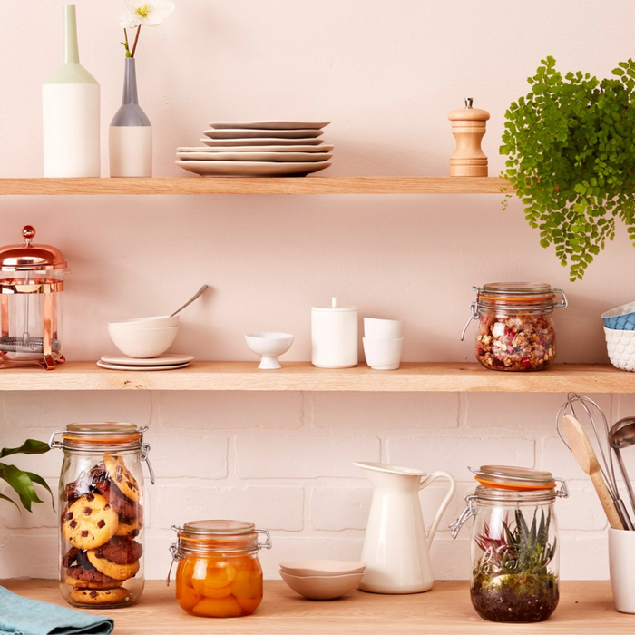 A kitchen scene shows wooden shelves with plates, cups, Le Parfait Super Jar Variety Pack with airtight seals filled with grains and plants. The countertop features canning jars with cookies and peaches, cutting tools, a white pitcher, and a small bowl against a light pink wall.