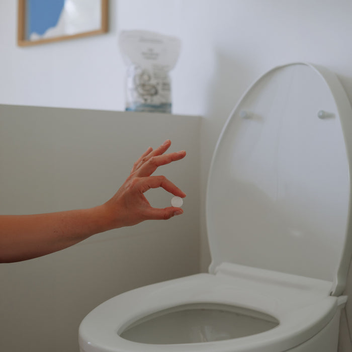 A hand holds The Unscented Company Toilet Bowl Cleaning Tab above an open toilet, ready to drop it in. In the background, a product packet and framed photo emphasize sustainable hygiene.
