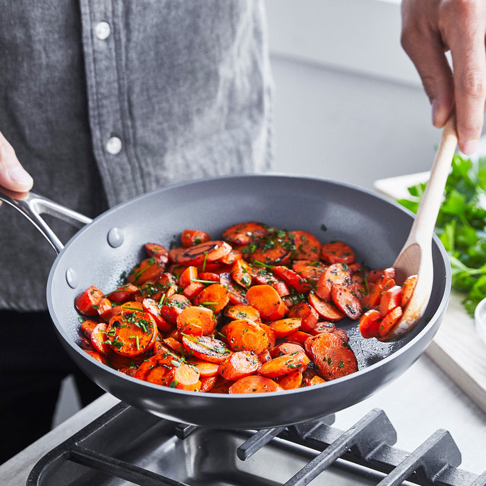 Someone in a gray shirt cooks herbed carrots in a GreenPan Valencia Pro Ceramic, Nonstick, Hard Anodized 12 Fry Pan, stirring with a wooden spoon. Parsley is visible in the background.