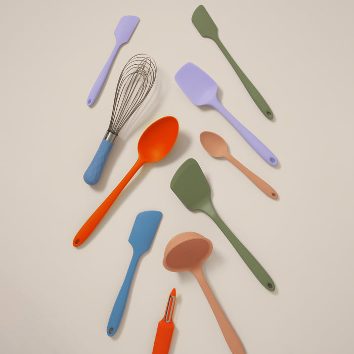 A 10-Piece Best Sellers Kitchen Tool Set by GIR is arranged on a beige surface, showcasing vibrant platinum-cured silicone kitchen utensils in orange, green, blue, and purple. It includes heat-resistant spatulas, a whisk, peeler, ladle, and spoon.