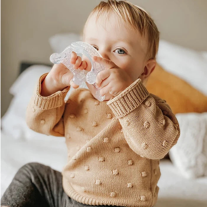 A toddler in a beige sweater with textured dots sits on a bed, holding Haakaa’s 100% Food Grade Silicone Sensory Development Crown Teether, a BPA-free item perfect for soothing gums. The child’s light hair contrasts softly against white and mustard-colored pillows.