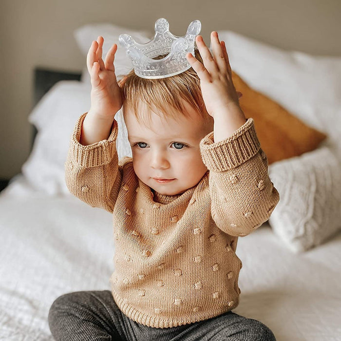 A toddler with light brown hair sits on a bed, wearing dark pants and a beige sweater, playfully holding the Haakaa 100% Food Grade Silicone Sensory Development Crown Teether above their head like royalty. This BPA-free toy is perfect for sensory development amid white and orange pillows.